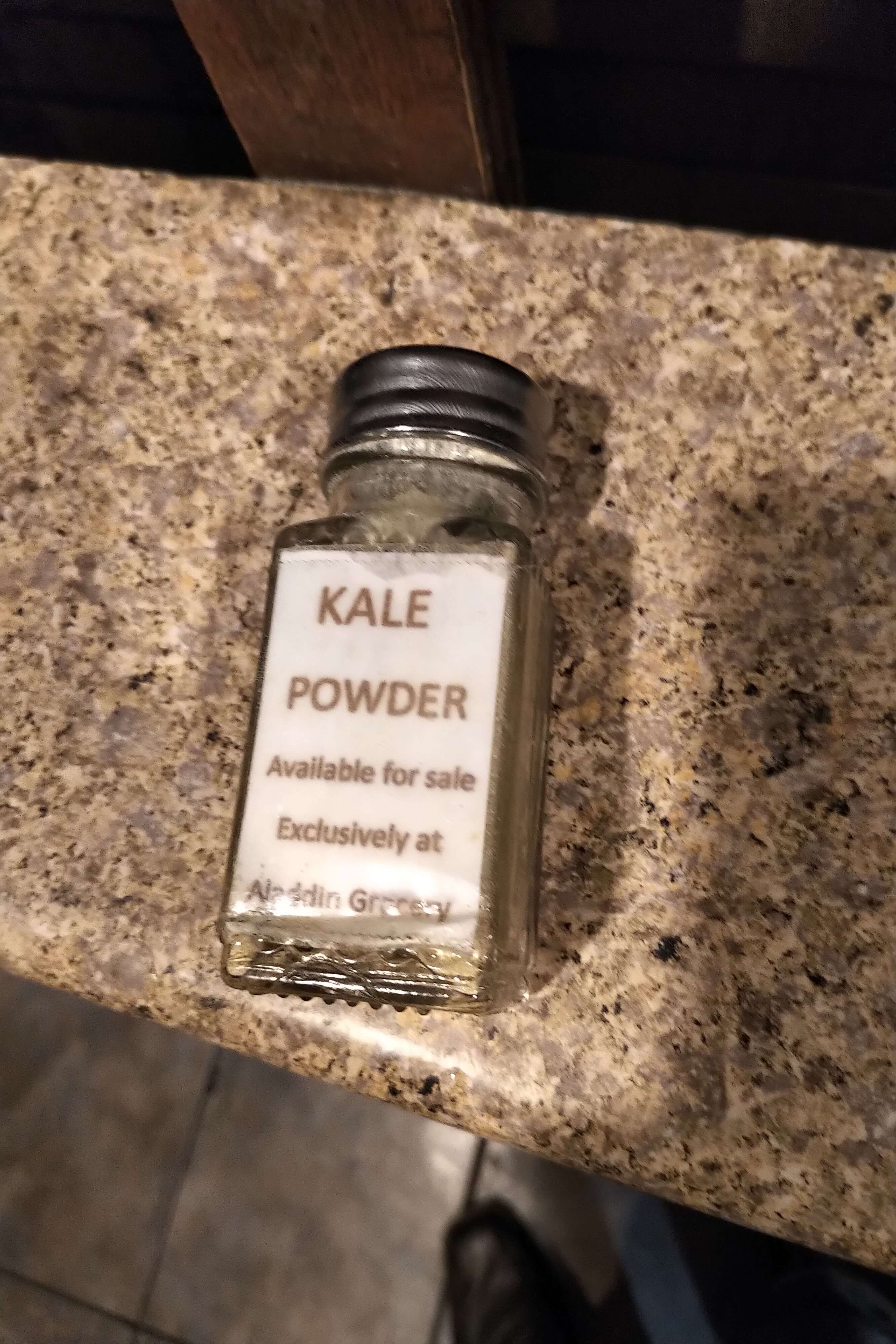 Old version of EasyKale kale shaker, resembling a glass salt shaker, with its name written on a paper label stuck to the container, displayed on a kitchen countertop.