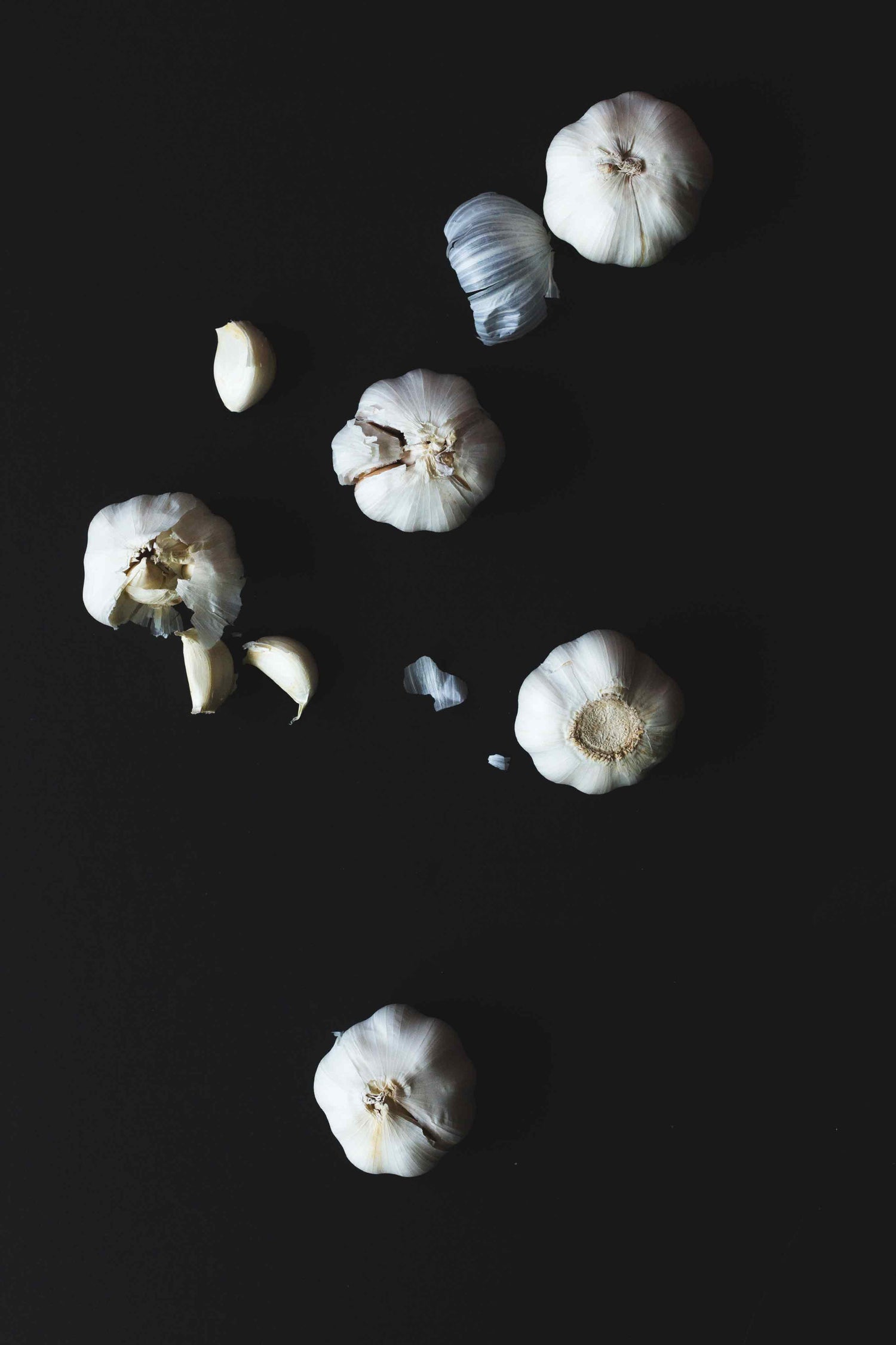 Cloves of garlic falling and bursting open, with bulbs spilling out, against a pitch black background.