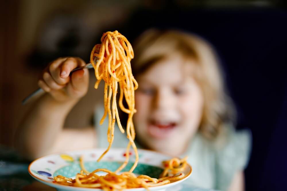 Close-up of a young girl's hands holding a forkful of spaghetti from a bowl, with a joyful smile in the background.